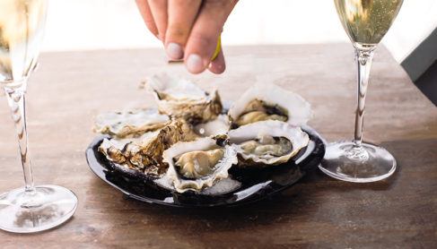 hand squeezing lemon on an oyster with white two glass of white wine on either side of the plate