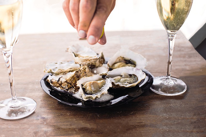 hand squeezing lemon on an oyster with white two glass of white wine on either side of the plate