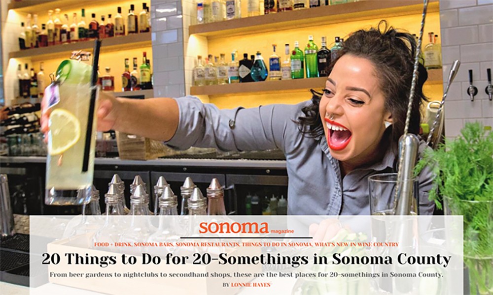 A happy and smiling female bartender at Perch + Plow handing a handcrafted cocktail to a customer with text overlay "Sonoma Magazine 20 Things to do for 20-something in Sonoma County"